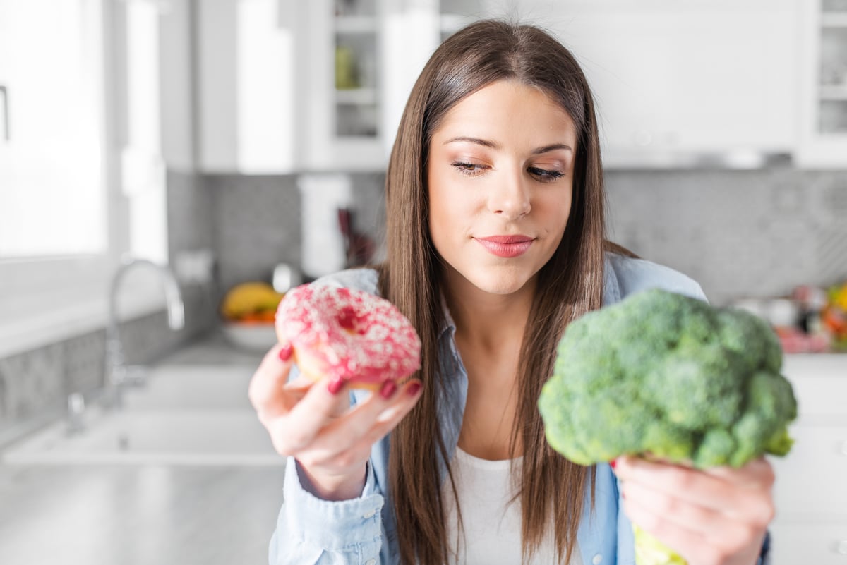 A person in a kitchen decides between eating a doughnut and broccoli.