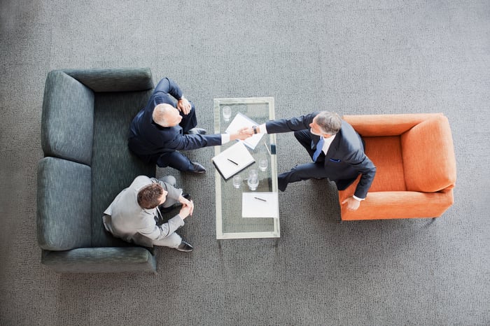 Three individuals sit at lounge chairs and shake hands.