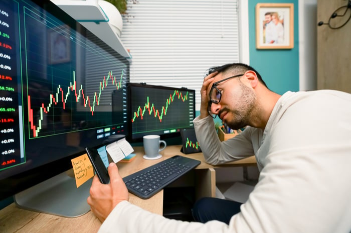 An investor touches their head in exasperation while looking at a phone and sitting in front of two screens displaying stock charts.