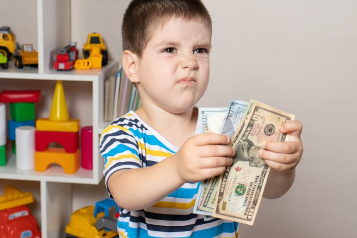 A child holds paper money in front of a shelf with toys and makes a disgusted face.