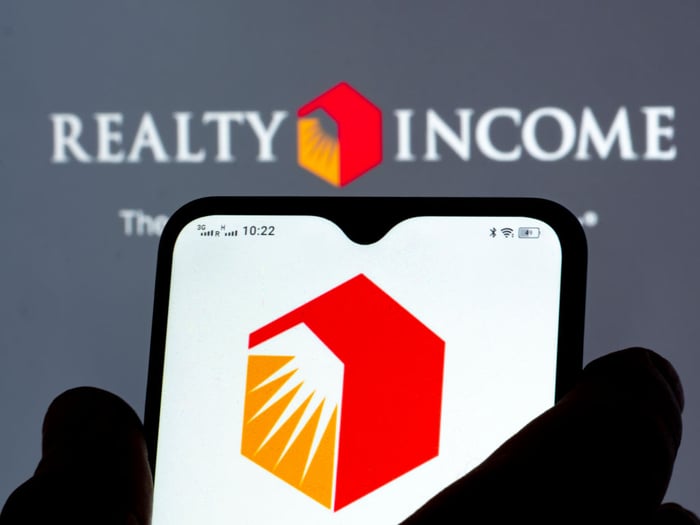 Realty Income logo on a smart phone with Realty Income's name and its logo on a wall in the background.