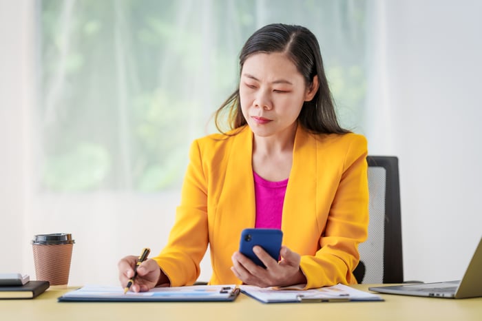 An investor sits at a desk while writing into a notebook and looking at a cell phone.