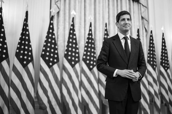 Kevin Warsh standing in front of a row of American flags in the East Room of the White House.