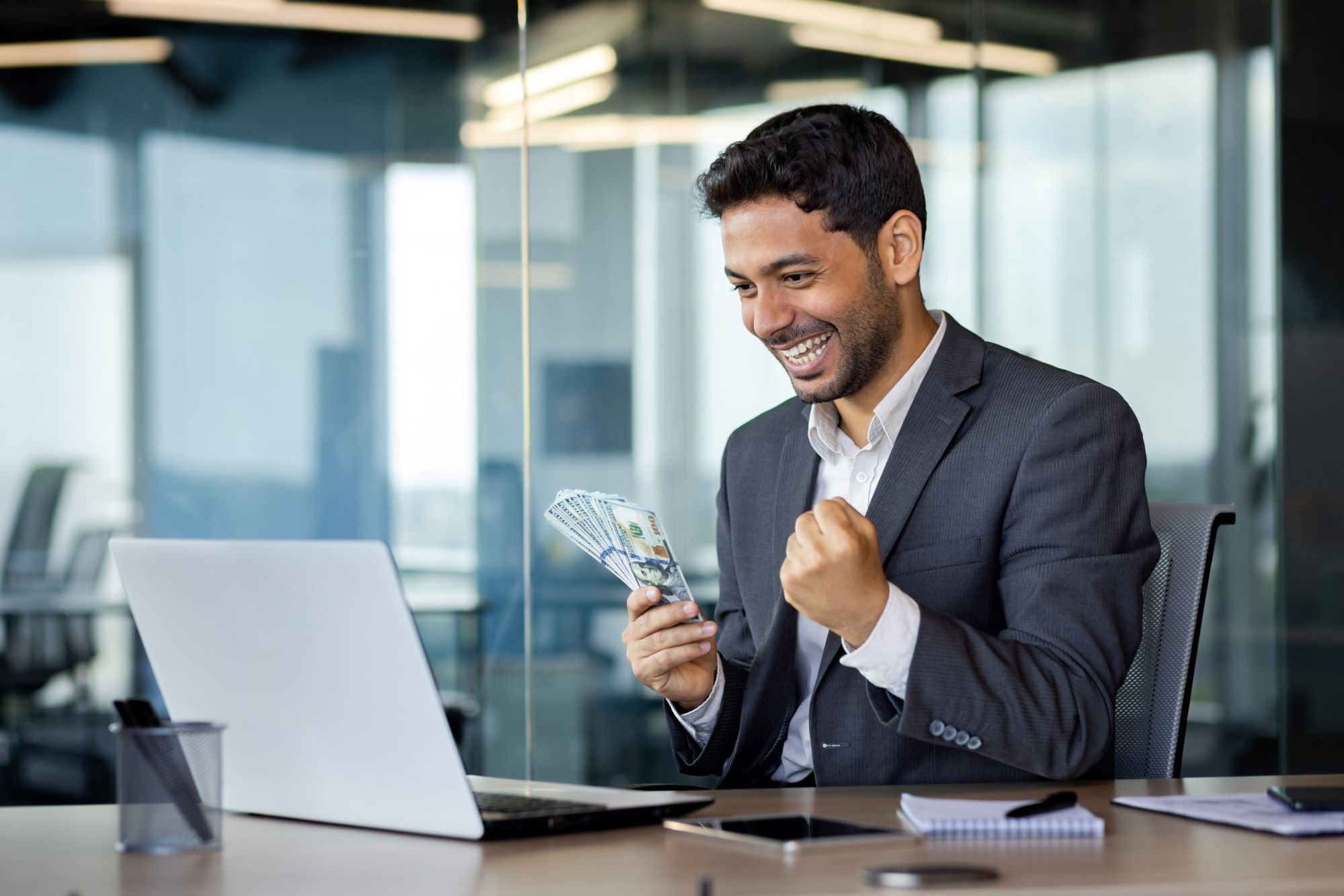 A happy person holding money in front of their computer.