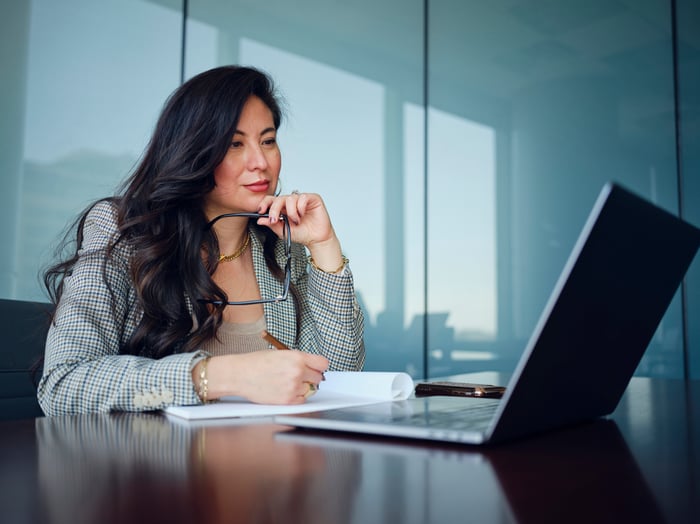 An investor sitting at a desk ponders a laptop while writing into a notebook.