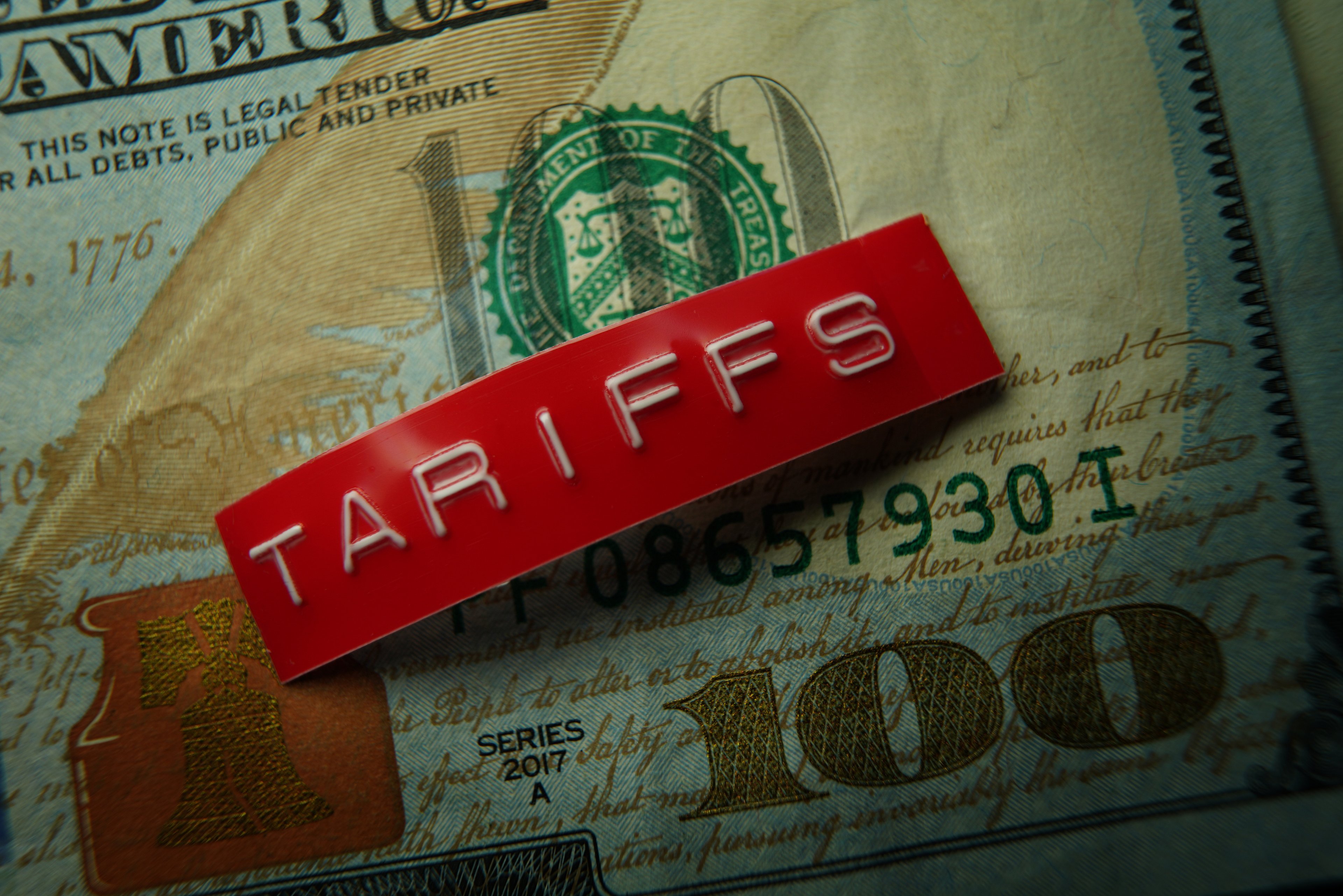 A stamped red metal badge that reads, tariffs, set atop a crisp one hundred dollar bill.