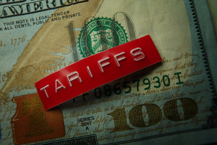 A stamped red metal badge that reads, tariffs, set atop a crisp one hundred dollar bill.
