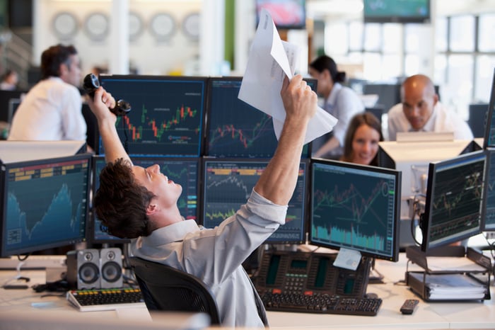 A trader sitting in front of six monitors is waving hands excitedly in the air.