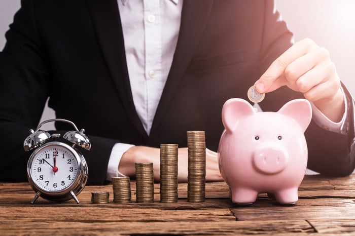 A person drops a coin inside a piggy bank, beside which is an alarm clock and stacks of coins arranged in an ascending pattern.