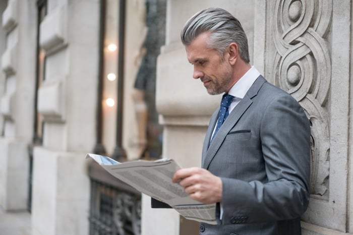 An investor wearing a gray suit reads from a newspaper while leaning against the stone facade of a building.