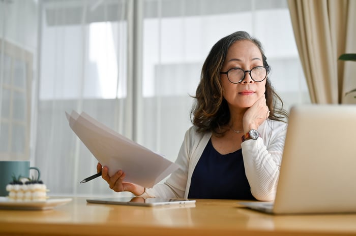 A businessperson holding paperwork in their right hand while reading content from an open laptop. 