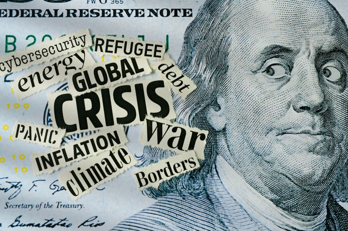 A $100 bill with Benjamin Franklin's eyes looking sideways at a group of newspaper clippings of words like Crisis, Debt, and Inflation.