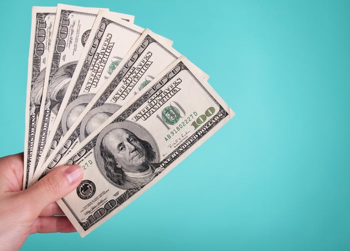 A person's left hand holding five fanned-out $100 bills.