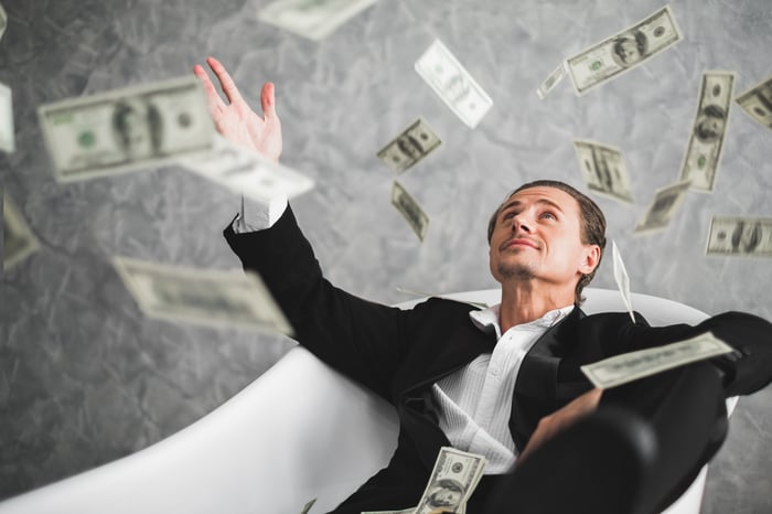 Man in a suit sitting in a bath tub amid flying currency notes.