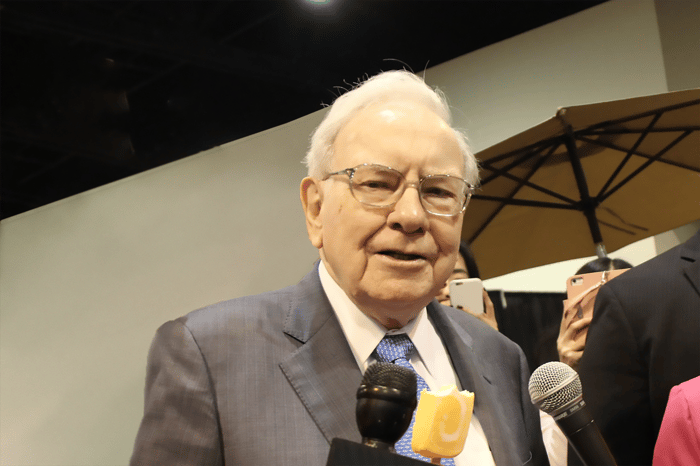 Warren Buffett eating an orange Popsicle with a microphone in front of his face.