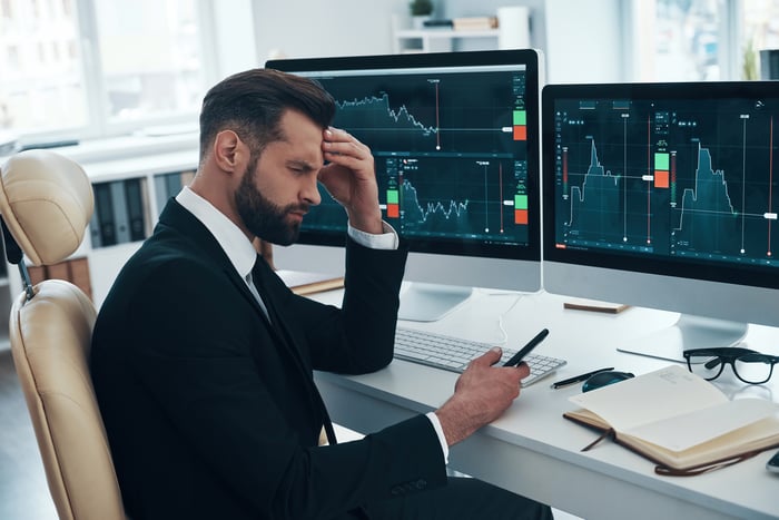 A person touches their head in frustration while sitting in front of a computer displaying stock price data and looking at a phone.