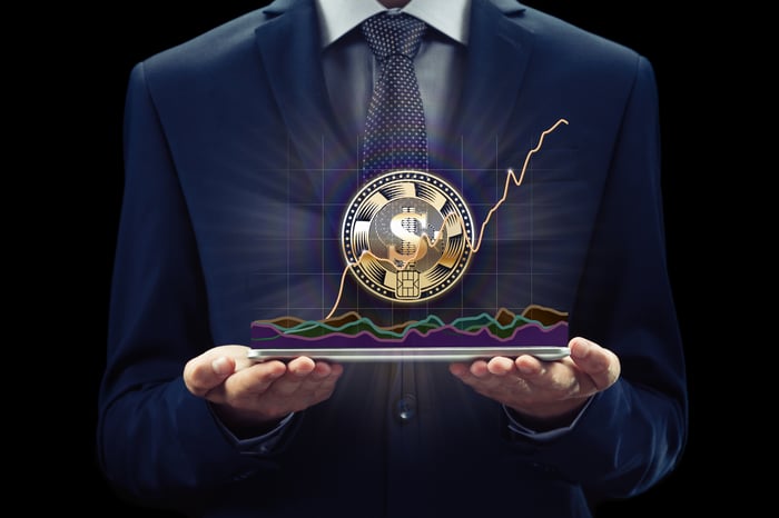 An investor holds a tablet underneath a cryptocurrency with a dollar sign that is floating in the air. 
