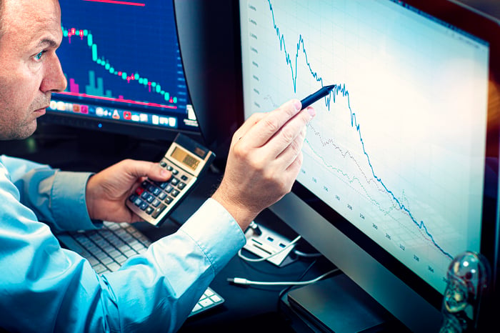 An analyst using a stylus and calculator to analyze a declining stock chart displayed on a computer monitor.