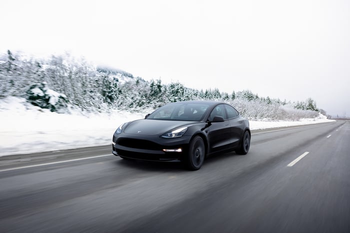 An all-electric Tesla Model 3 sedan driving on a two-lane road during wintry conditions.