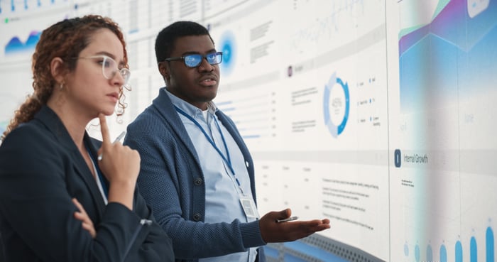 One analyst is speaking to another in front of a presentation screen.
