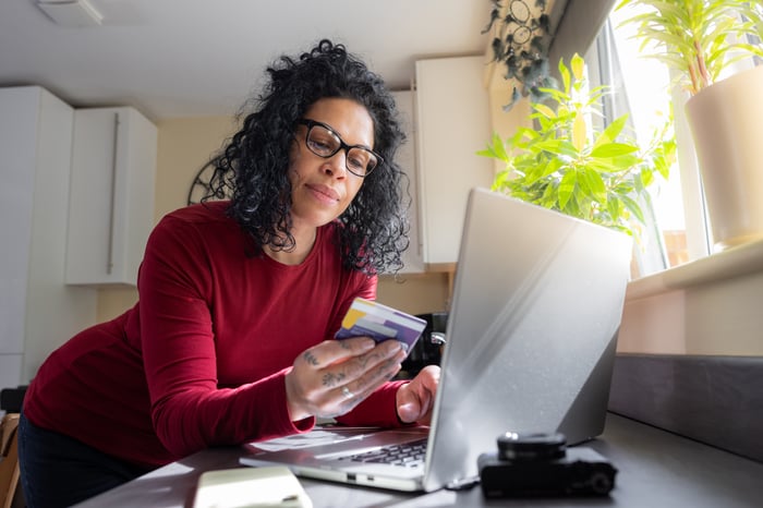 A person leaning over desk with laptop, looking at bank card. 