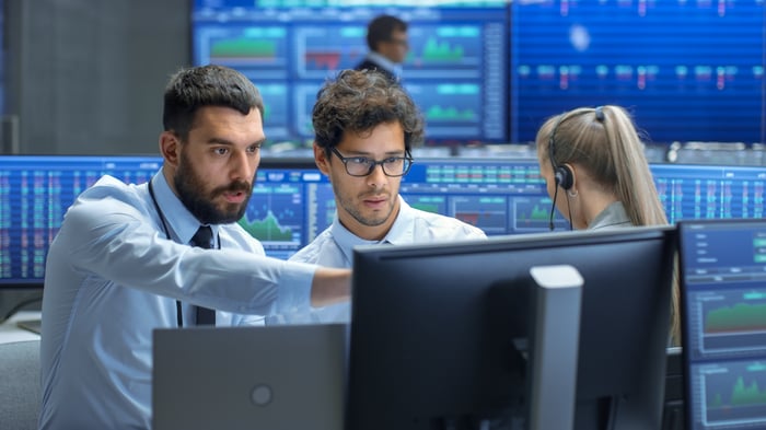 Two men looking at screens with other monitors showing financial data in the background. 