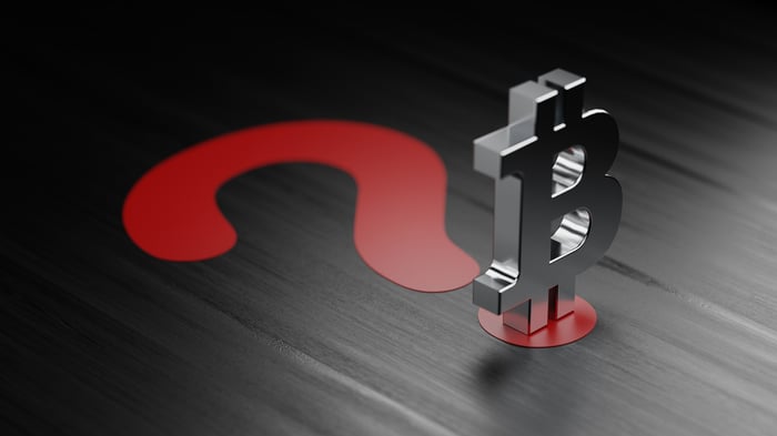 A metal Bitcoin logo figurine atop a large, red question mark.