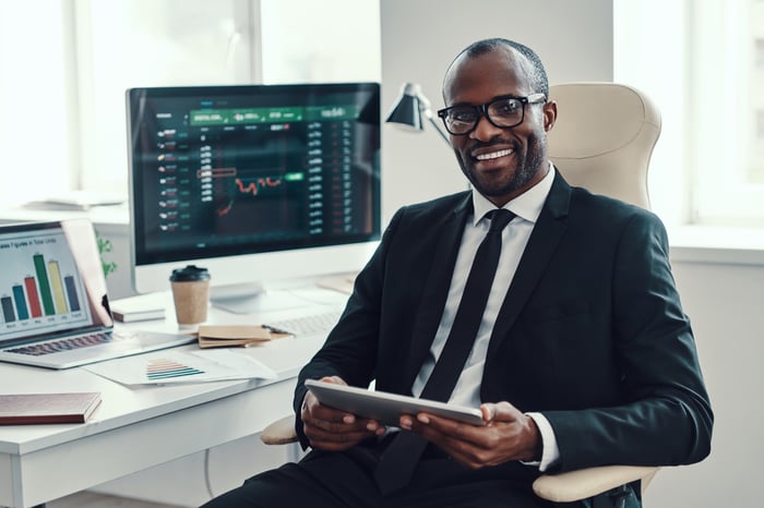 Smiling person at desk, beside computers displaying charts.