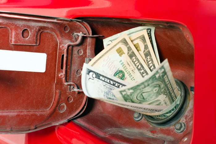 Cash bills of various denominations sticking out of a red car's gas tank.