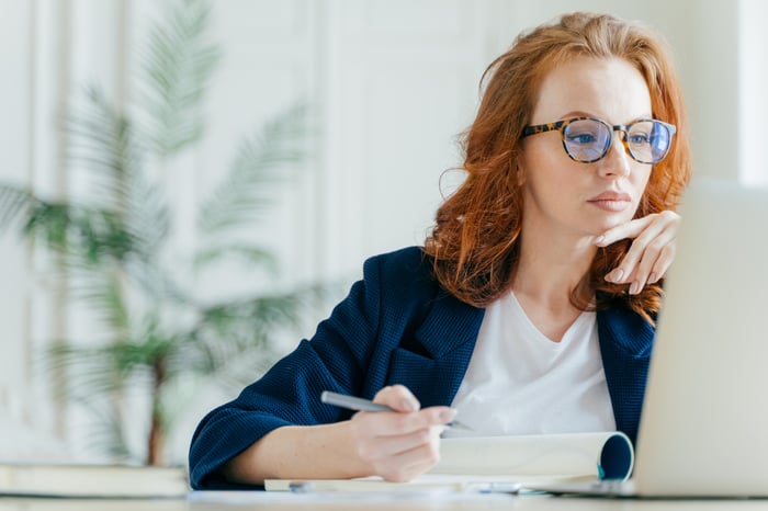 Person sitting at desk, looking at laptop and taking notes.