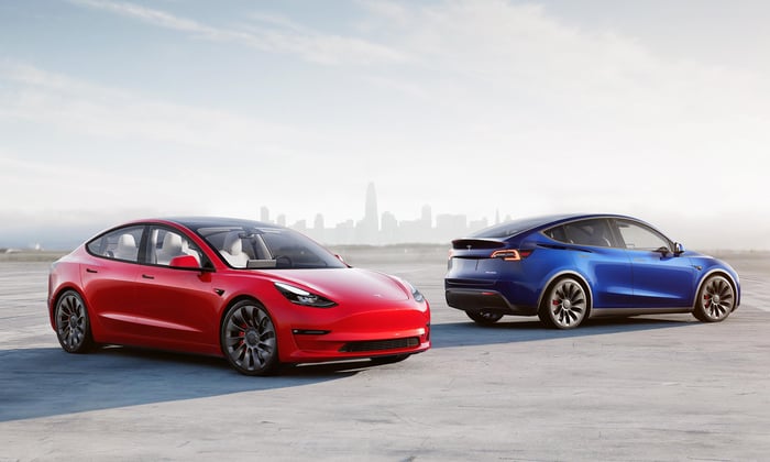 Two Tesla EVs parked on a wide, paved surface with a metropolitan skyline faintly seen in the background. 