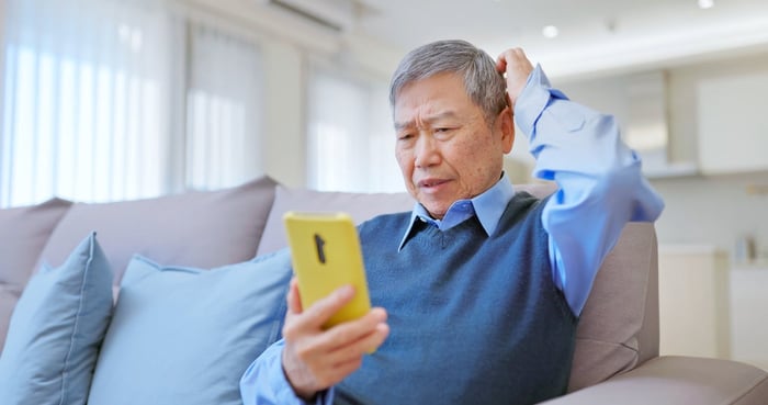 Older gentleman sitting on a sofa, looking at his smartphone with a confused expression on his face. 