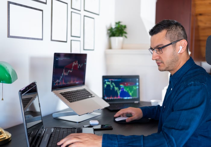 An investor consults three laptop computers displaying stock price data while sitting at a desk.