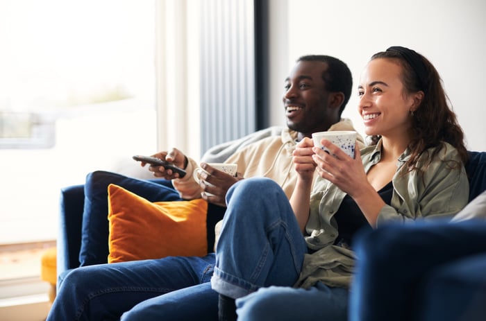 Two people channel surfing from a couch.