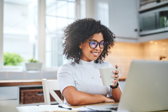 An investor holds a cup while studying something on a laptop at home.