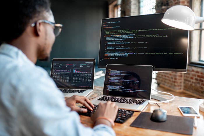 Man in specs working on a laptop with two additional screens behind. 