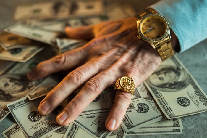 An older person's hand with gold watch and pinky ring on a pile of hundred-dollar bills.