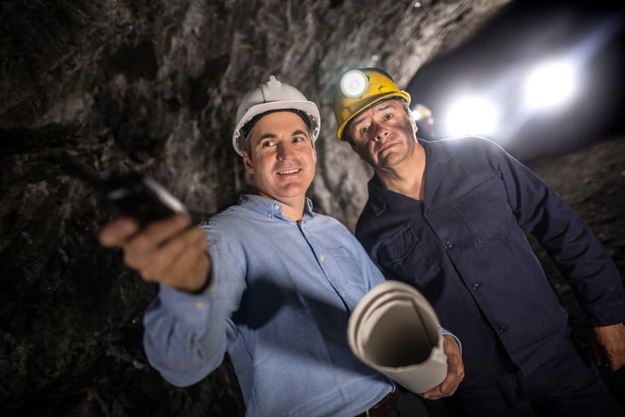 A miner and an engineer wearing hard hats are in a mine shaft with lighting behind them.