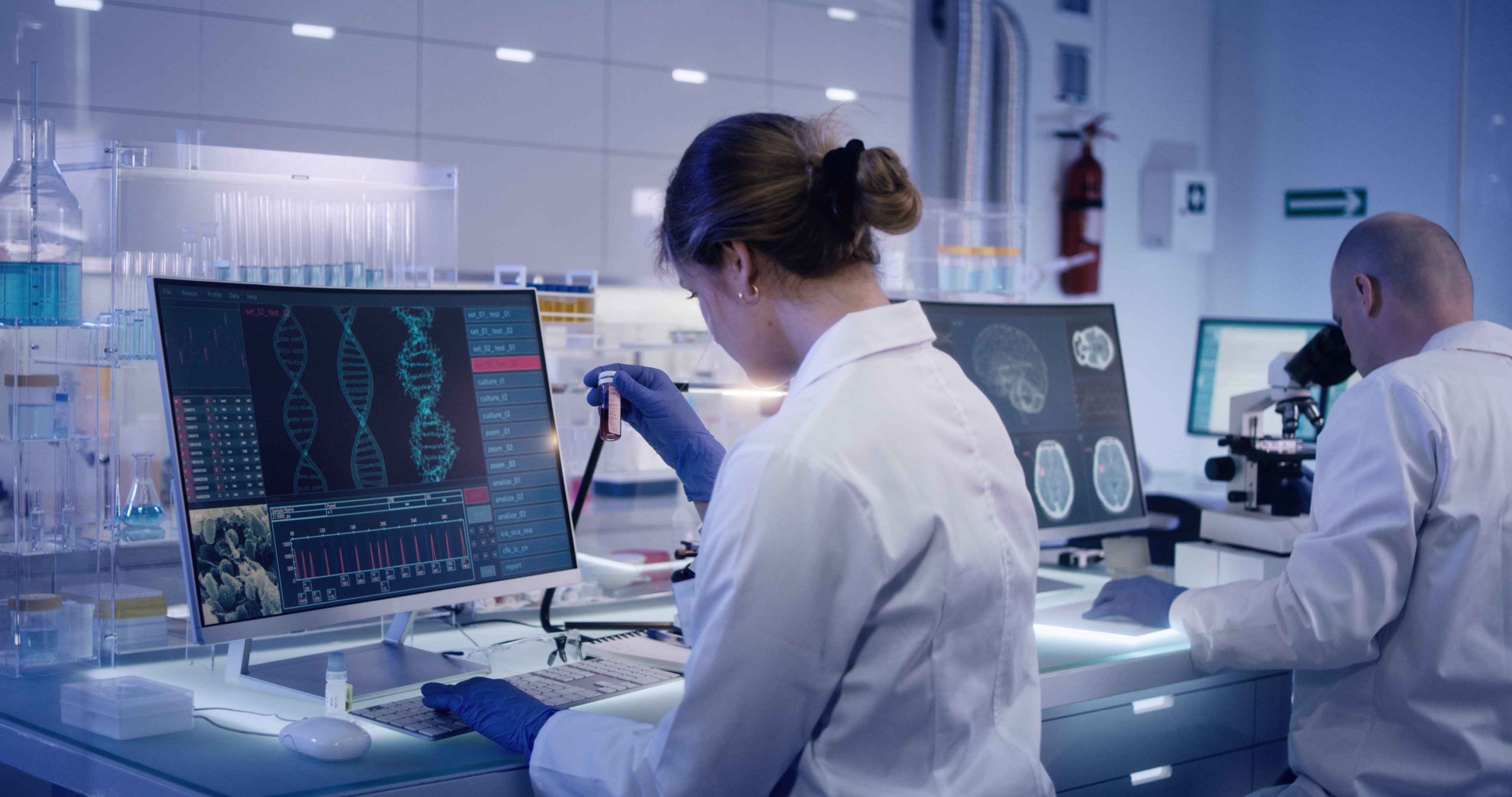 Scientist in lab coat studies a test tube of fluid while looking at a strand of DNA on a computer screen