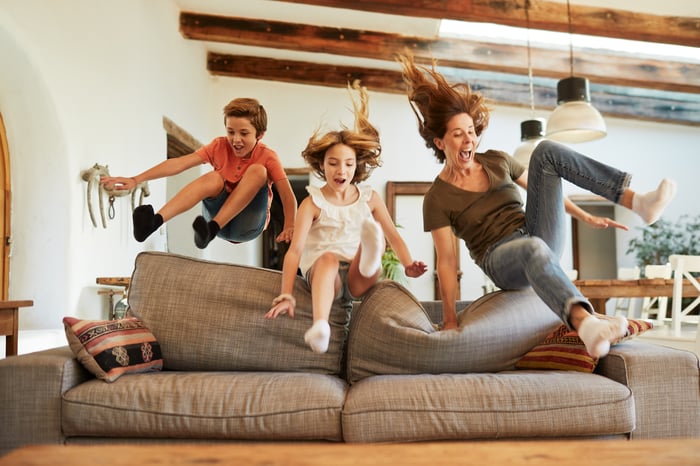 A mom and two kids jumping onto a couch.
