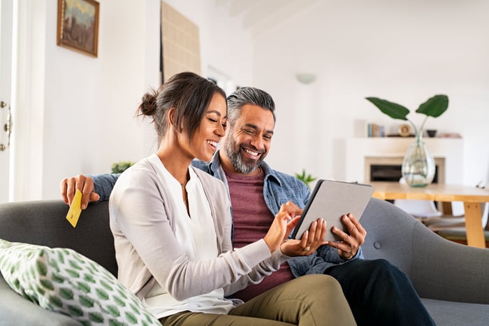 Two investors sit on a couch in a living room and look at something on a tablet. 