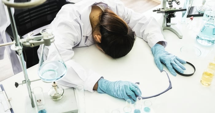 A person wearing a white lab coat and rubber gloves with head down on a table and lab equipment near the person.