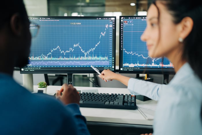 A person points to a screen with stock market charts, with another person sitting next to them.