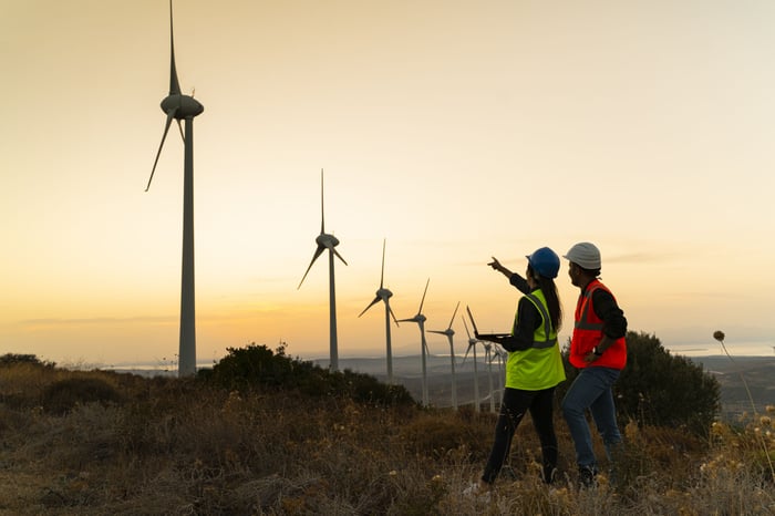 Two engineers are assessing a long line of wind turbines as the sun is setting.