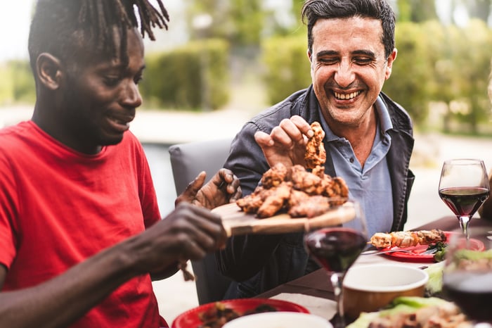 Two people laugh while sharing a plate of chicken wings and red wine at an outdoor table.