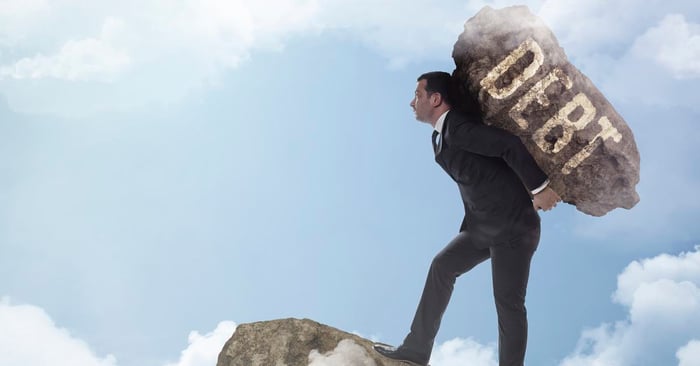 Man in suit carrying boulder labeled DEBT to edge of cloudy precipice.