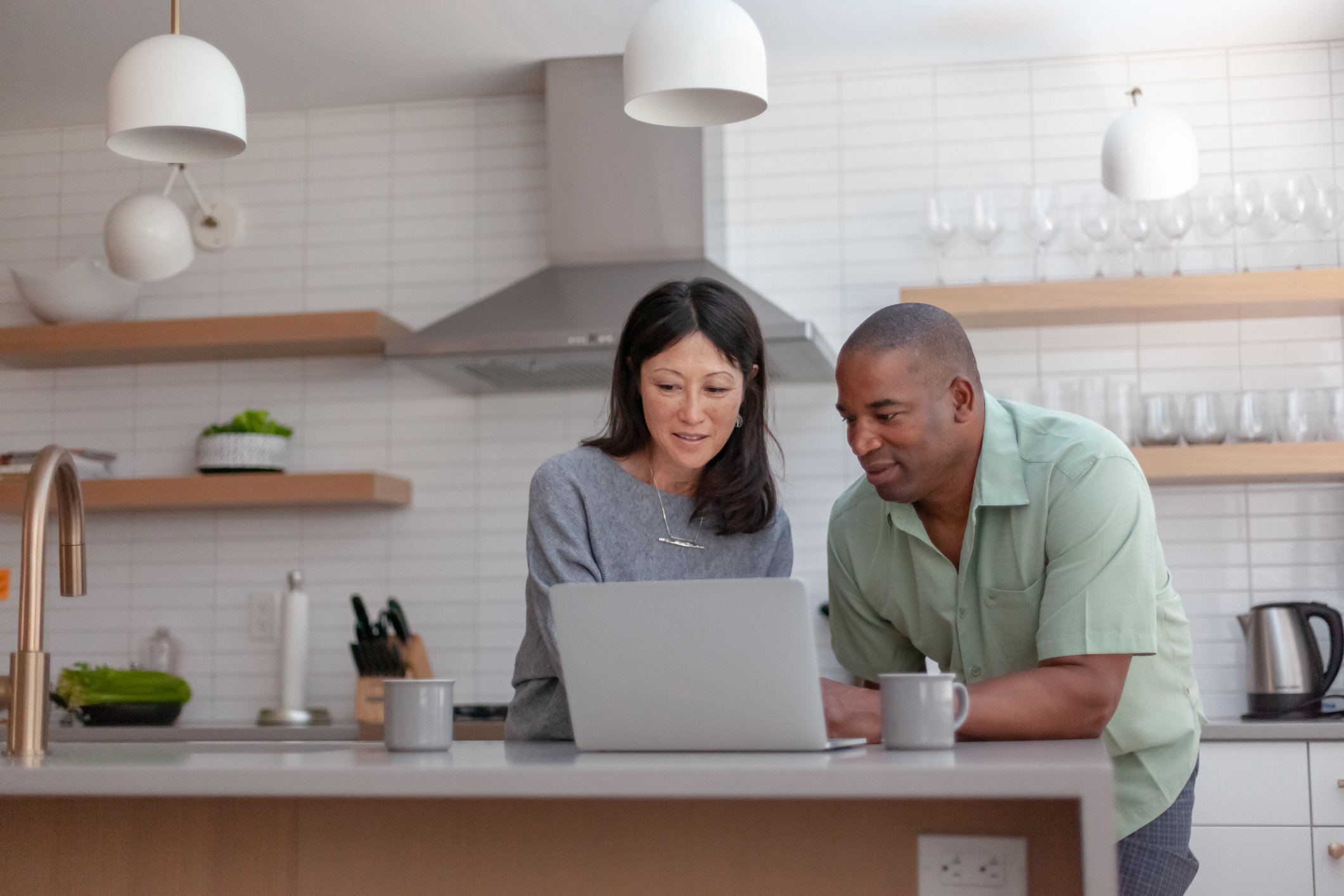 Two people talk while looking at laptop in kitchen.