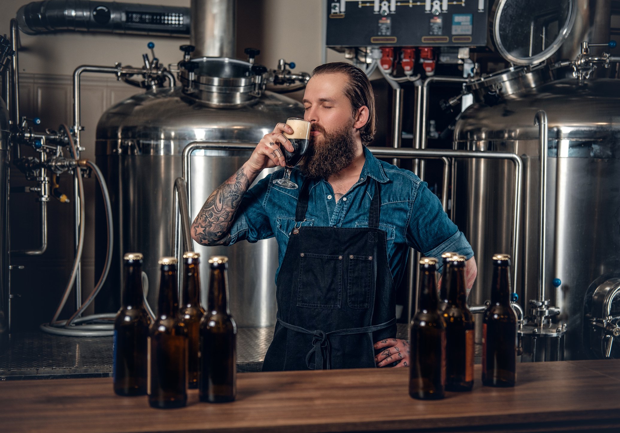 Man with a beard stands inside brewery holding a glass of beer near his nose and mouth.