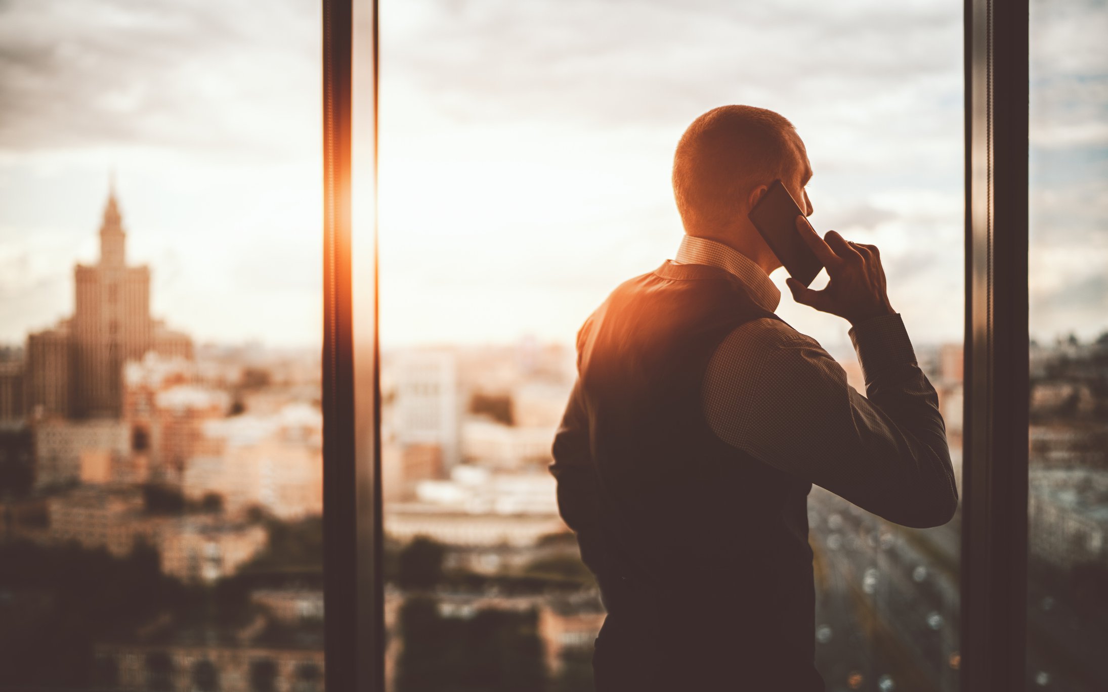 Businessperson standing at office window while looking out at the city and talking on the phone