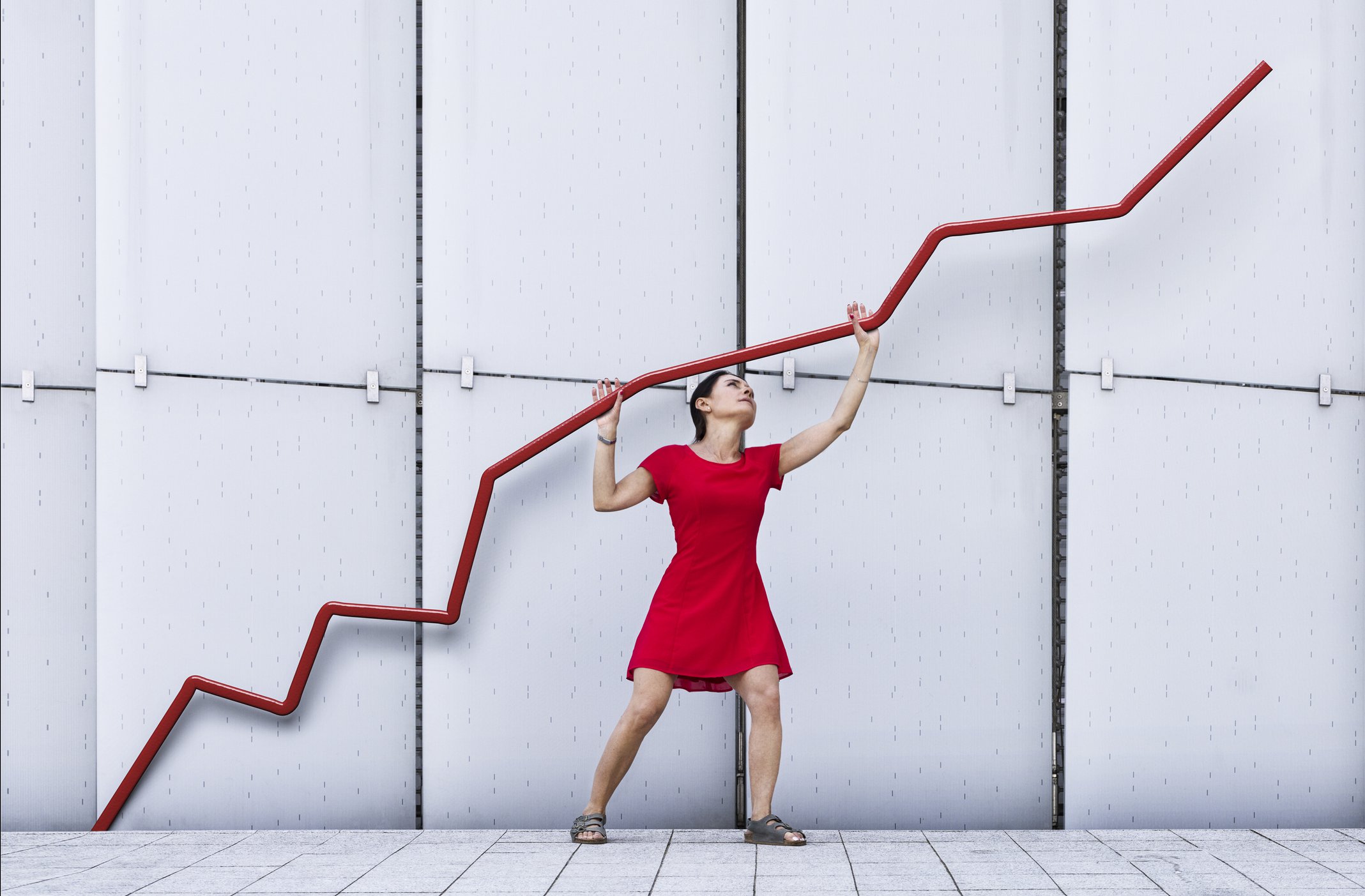 Woman in red dress propping up red trend line going upward against white wall